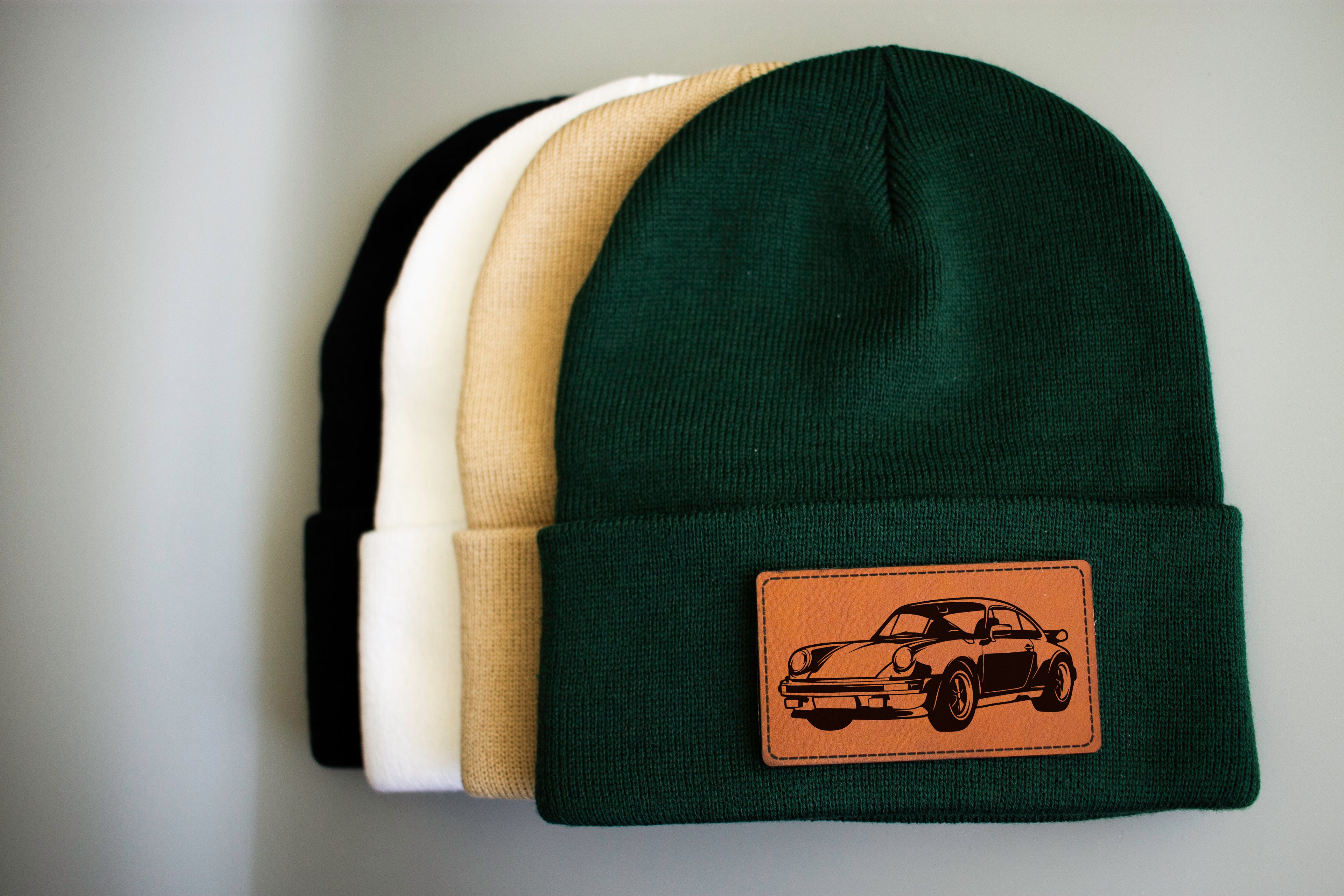 Four beanie hats in black, white, beige, and green with a leather patch featuring a car design on a gray background.
